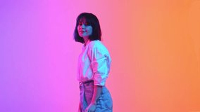 Young brunette woman posing in casual attire and looking at camera with smile in neon light against gradient studio background. Concept of human emotions, fashion, beauty, style. ad
