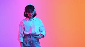 Young woman in oversize outfit listening music in headphones and dancing in neon light against gradient studio background. Concept of human emotions, fashion and beauty, style. ad