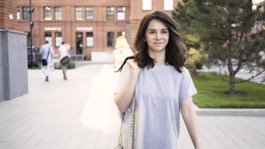 Cheerful young woman with dark hair wearing a gray T shirt is walking from a clothes store with paper bags and waving to a friend off screen. Tracking real time medium shot
