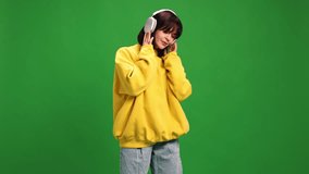 Young woman in modern oversize outfit listening music in headphones and dancing against green studio background. Concept of human emotions, fashion and beauty, style, technology. ad