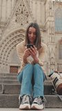 Vertical video, Happy traveler girl using mobile phone while sitting on the steps of an old building in the historic part of an old European city