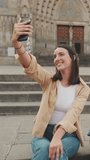 Vertical video, Traveler girl taking selfie on mobile phone while sitting on the steps of an old building in the historical part of an old European city