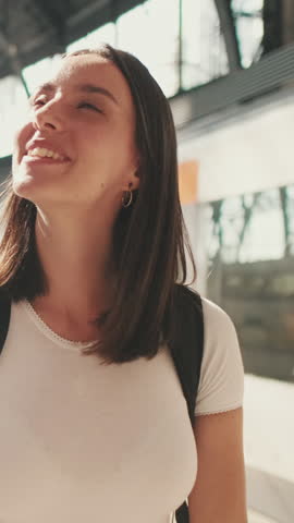 Vertical video, Beautiful traveler girl with backpack on her shoulders walks along the platform of the railway station in her hands with mobile phone