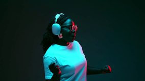 Relaxed African-American woman listening music in headphones and singing in red neon light against gradient studio background. Concept of human emotions, beauty and fashion, style,