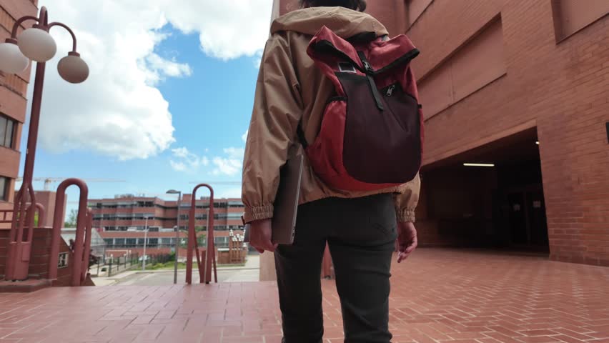 A Latin-American woman strolls through the university campus with her laptop and backpack on a sunny day, heading to classes. University student life, education, and future