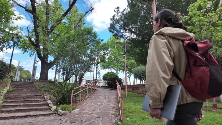 A Latin-American woman strolls through the university campus with her laptop and backpack on a sunny day, heading to classes. University student life, education, and future