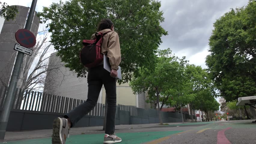 A Latin-American woman strolls through the university campus with her laptop and backpack on a sunny day, heading to classes. University student life, education, and future