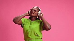 Relaxed African-American woman listening music in headphones and singing against pastel pink studio background. Concept of human emotions, fashion and beauty, trends, party, modern lifestyle.