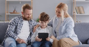 Happy young parents with little child boy sitting on sofa using modern tablet together for social media or watching funny cartoon enjoying weekend. Family leisure concept. 4k video.