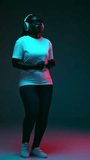 Overjoyed African-American woman listening music in headphones and dancing in red neon light against gradient studio background. Concept of human emotions, beauty and fashion, style,
