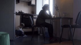 Worried mother sits at kitchen table, contemplating, as child plays quietly behind her. Domestic life with subtle tension, family issues, indoor, natural light. - Powered by Shutterstock - Get 15% off with code: PIKWIZARD15