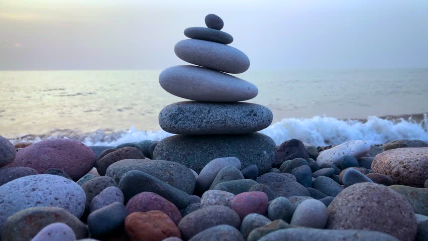 Balanced sea stones on a pebble beach with foaming and waving sea. Balanced pebble stones pyramid close up. Calm, harmony, zen, spa, relaxing meditation background. Sea water waves crashing at sunset.