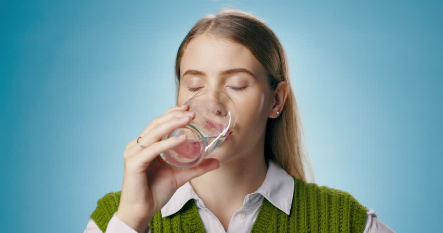 Woman, drinking water and glass in studio to hydrate on break for gut health, diet or detox by blue background. Girl, university student or person for wellness, aqua or vision for nutrition in London