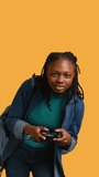 Vertical video African american gamer holding controller, focused on finishing difficult videogame level. Woman gaming with gamepad, solving quests, isolated over studio backdrop, camera B