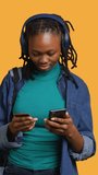Vertical video African american teenager adding payment method on website, isolated over studio background. Young girl wearing headphones, doing internet shopping, typing credit card information on