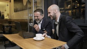 Two charismatic middle-aged businessmen, dressed in suits, are having a meeting outside the office in a cafe environment, laughing heartily while working with their laptops - Powered by Shutterstock - Get 15% off with code: PIKWIZARD15