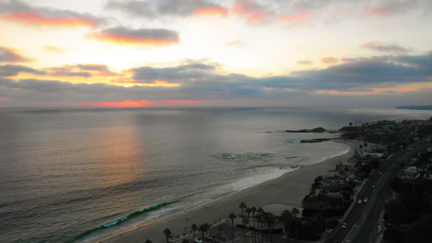 Aerial Panning Backward Scenic View Of Cars Moving On Road By Sea At Tranquil Beach Under Cloudy Sky At Sunset - Laguna Beach, California