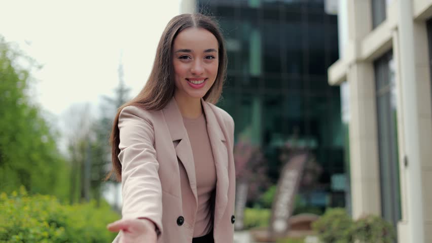 Attractive female smiling at camera and making inviting gesture with extended hands on city street background. Friendly lady spinning around and asking to follow in exploring new spot around.