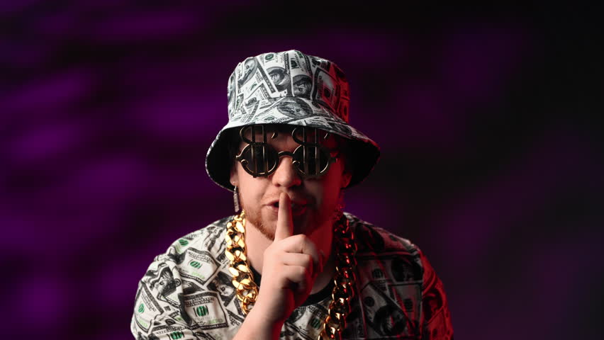 Crazy rich man showing sign of silence and making shh, silence gesture with finger, portrait in studio. Extravagant person in clothes with dollars symbol asking to be quiet and save secret of wealth