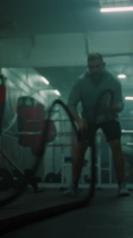 Athletic man exercises with battle ropes in dark boxing gym. Professional male boxer does cardio or endurance workout before championship fight. Physical activity Vertical shot