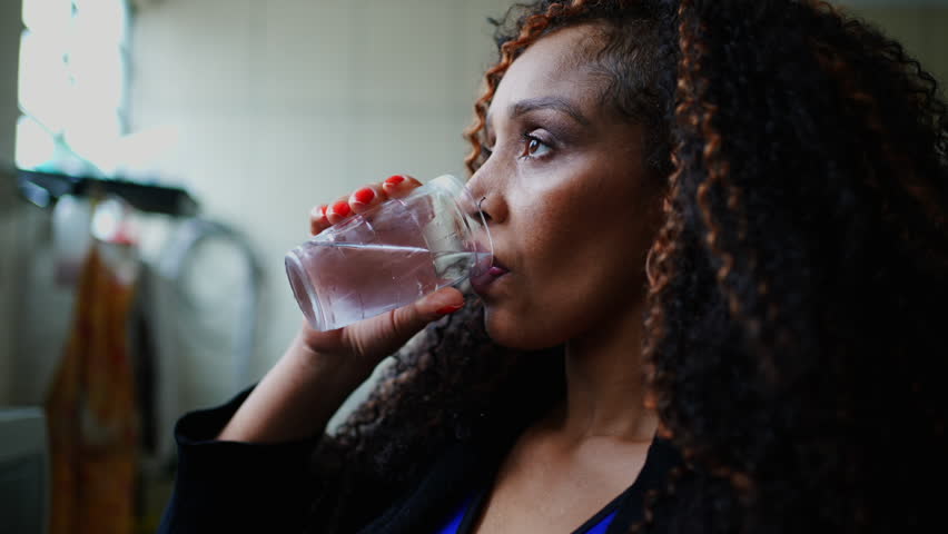 One thoughtful black middle-aged woman drinking water while gazing in the distance with pensive emotion. Contemplative latina person of African descent hydrating herself