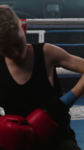 Vertical shot Teenager sits near boxing ring and puts on boxing gloves. Young boxer comes in boxing ring and starts practice punches with African American trainer. Athlete prepares to competition in