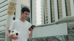Asian Teenager Walking Home And Browsing Internet Holding The Football Ball. Handsome Guy Soccer Player Going Outside And Scrolling Apps After Football Exercises. Football and Technology Concept - Powered by Shutterstock - Get 15% off with code: PIKWIZARD15