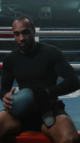 Vertical shot fighter sits near ring and puts on boxing gloves. Adult boxer comes in boxing ring and starts to train punches with coach. Athlete prepares to tournament or competition in boxing gym.