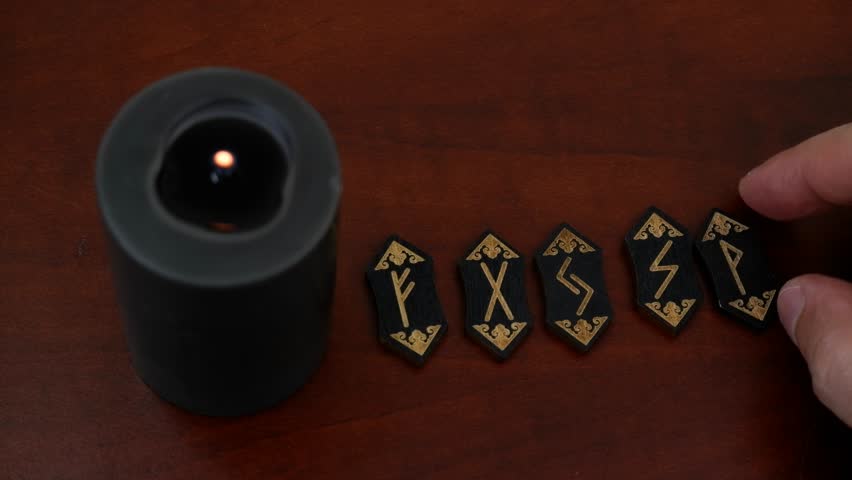 Burning black witch candle and black runes on wooden table.	Hand choosing different runes, fortune telling concept.