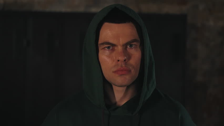 Focused boxer in green hoodie stares directly at camera from dimly lit locker room, exuding intensity and determination. Grit and readiness of fighter athlete moments before entering ring