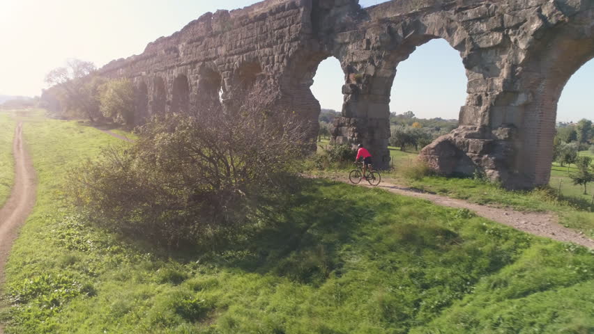 Young man cyclist on dirt road along ancient roman aqueduct in orange sportswear aerial view drone