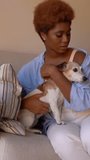 Happy smiling Black woman petting small white dog sitting on sofa in living room. Dog is kissing licking. weekend leisure time with pet at home. Vertical video footage