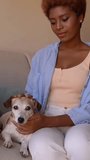 Black woman petting small white dog sitting on sofa in living room. Caring touches of dog owner. weekend leisure time with pet at home. Vertical video footage
