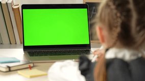 A cute little girl using with a laptop. Green screen for replacement. School girl looks at a laptop with mockup green screen. Distance education concept.