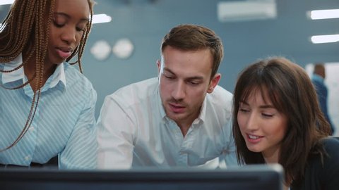 Three people standing at computer. Beautiful girl with African pigtails pressing key on keyboard and turning on video presentation. Young people smiling, talking. Office. Close-up.