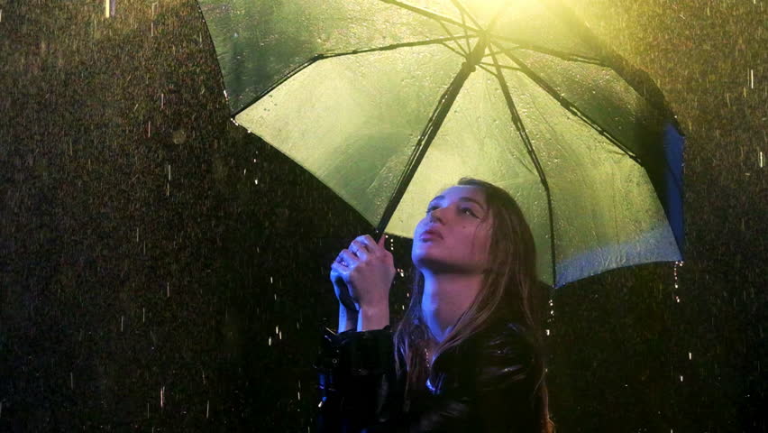 Seductive blonde woman stands in rain with umbrella. Keeping head high lady observes streaming water cascading onto wet coat