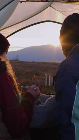 Multiethnic couple of hikers looks at sunset and talks. African American man sits in tent on top of hill and drinks tea with Caucasian woman. Young tourists rest in mountains on their vacation.