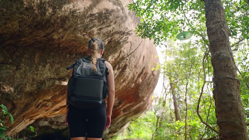 Active woman with backpack explores trail in rocky mountain grotto. Hiker adventure through natural cave landscape, sunlight filtering through trees. Solo traveler treks in wild, scenic nature. Slowmo