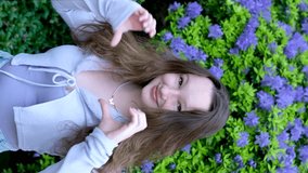 young girl shows a heart with her hands laughs strongly shakes her head shows a thumb eye. vertical video smile on the background of a blue bush of flowers