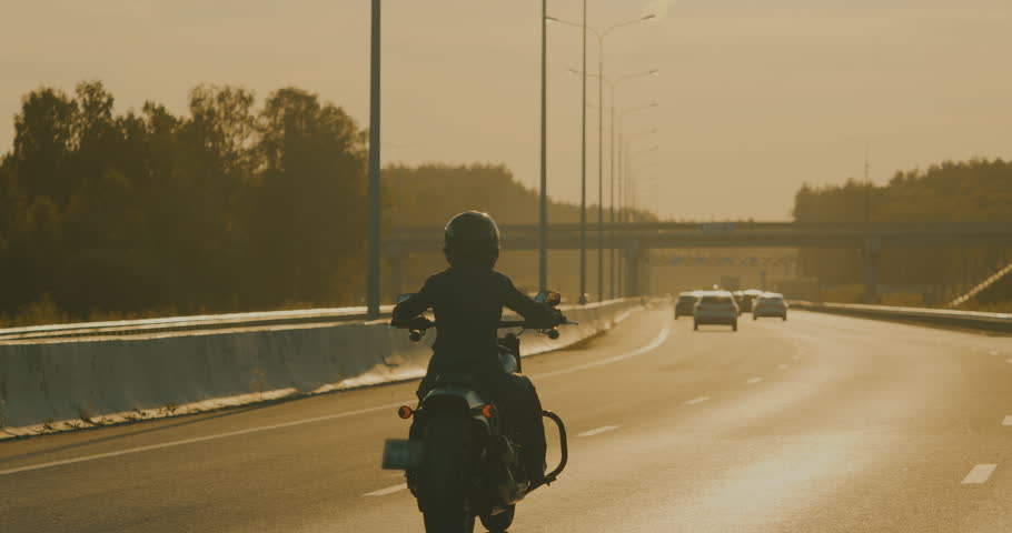 Brutal woman in helmet riding fast modern motorbike at highway. Motorcyclist racing his motorcycle on asphalt road. Driving bike during trip at sunset lighting, concept of adventure, back view