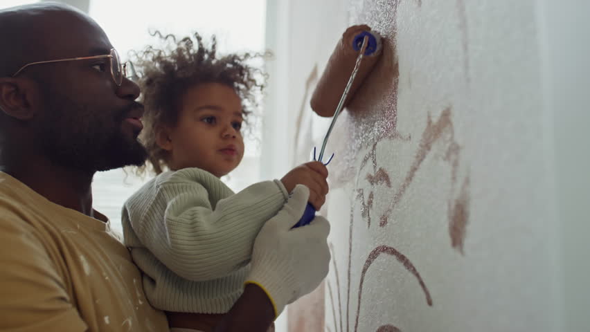Side closeup of positive African American dad holding his Biracial baby daughter in hands and helping her to paint wall in brown using roller Royalty-Free Stock Footage #3494259441