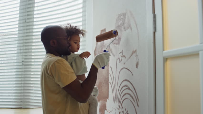 Side footage of African American man holding his little Biracial daughter in hands and teaching her how to paint walls with roller during room redecoration Royalty-Free Stock Footage #3494259619