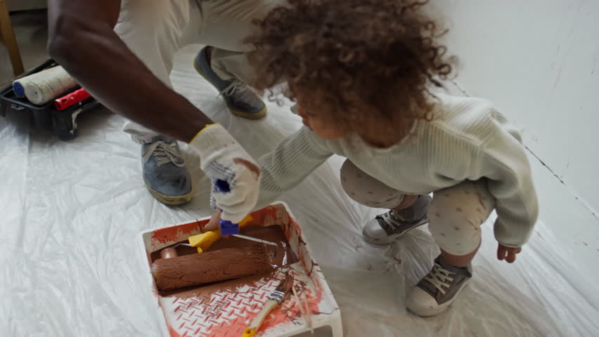 Side footage of little Biracial baby girl diving her mini paint roller into tray and painting walls in future nursery in brown, helping African American father during room renovation Royalty-Free Stock Footage #3494262581