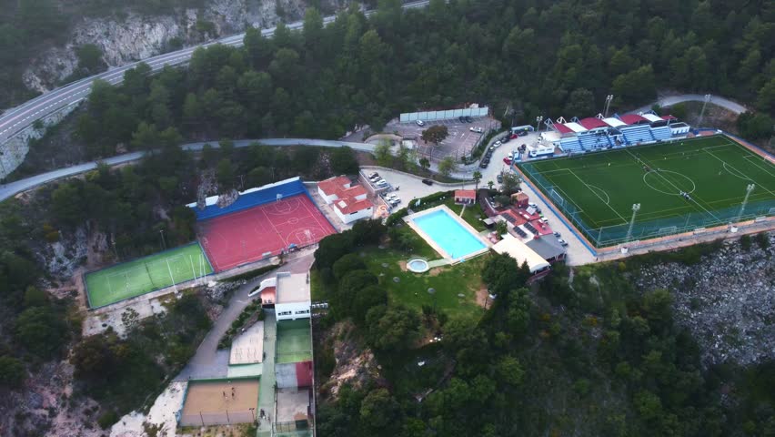 High-angle view of a sports complex nestled in a lush green area, featuring a football field, swimming pool, and tennis courts