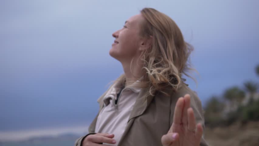 Happy female tourist in a long raincoat enjoying the view of the cloudy sky and the sea from the mountain.