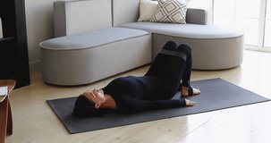 An athletic woman performs exercises to strengthen the muscles of the thighs with a fitness band with raising the body from a lying position, training on the mat in black sportswear, full-length view