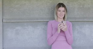 A young Caucasian woman dressed in a fitted pink sports outfit rests against a concrete wall, using her smartphone after a workout session. This clip perfectly illustrates the balance between fitness