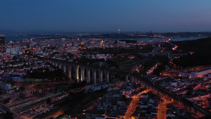 Illuminated Lisbon Skyline, Aguas Livres Aqueduct and 25th April Bridge at Night. Blue Hour. Portugal. Aerial View. Orbiting
