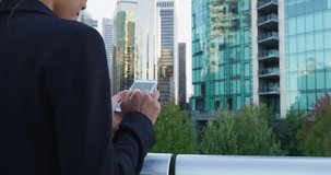 Young businesswoman using mobile phone against office buildings. Female business executive is text messaging through smart phone in financial district. She is wearing suit in city.