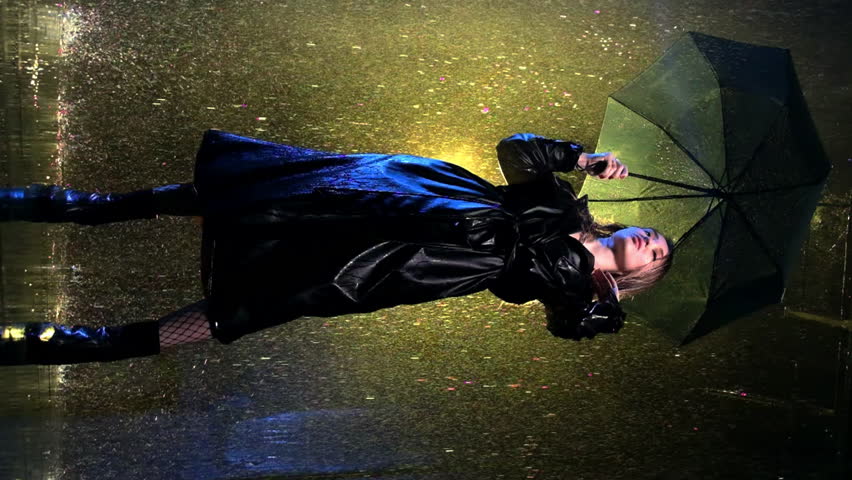 Woman stands amidst heavy rain holding black umbrella. Illuminated by street lamp lady wears alluring tights and leather trench coat